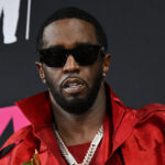 Sean "Diddy" Combs in a suit and sunglasses, looking to the side, with a blurred background of people and cameras, representing the media attention surrounding the case.