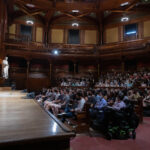 Man at podium on stage speaking to large group of students sitting in theater seating.