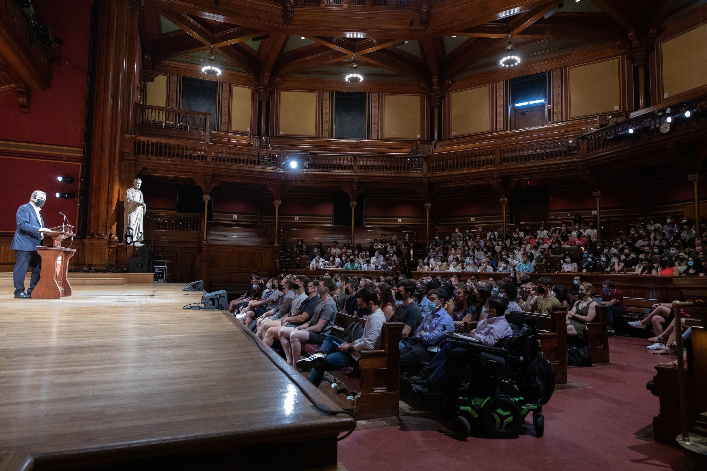 Man at podium on stage speaking to large group of students sitting in theater seating.