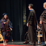 A student in a blue commencement gown and regalia, including a stole with the words