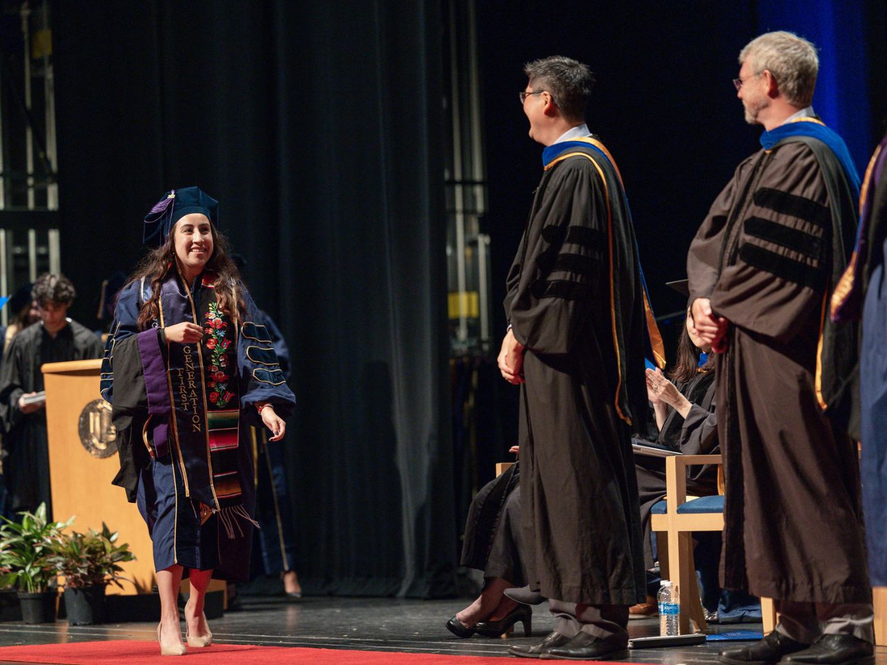 A student in a blue commencement gown and regalia, including a stole with the words